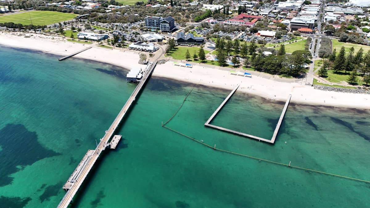 Luftaufnahme von Glenelg Beach in Adelaide mit dem Pier, dem Seebadebereich, klarem türkisfarbenem Wasser, sandigem Ufer und der nahegelegenen Stadt.