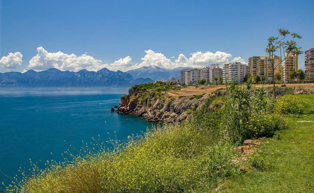 Clifftop park in Antalya with green grass, wildflowers, and buildings overlooking the Mediterranean Sea and distant mountains.