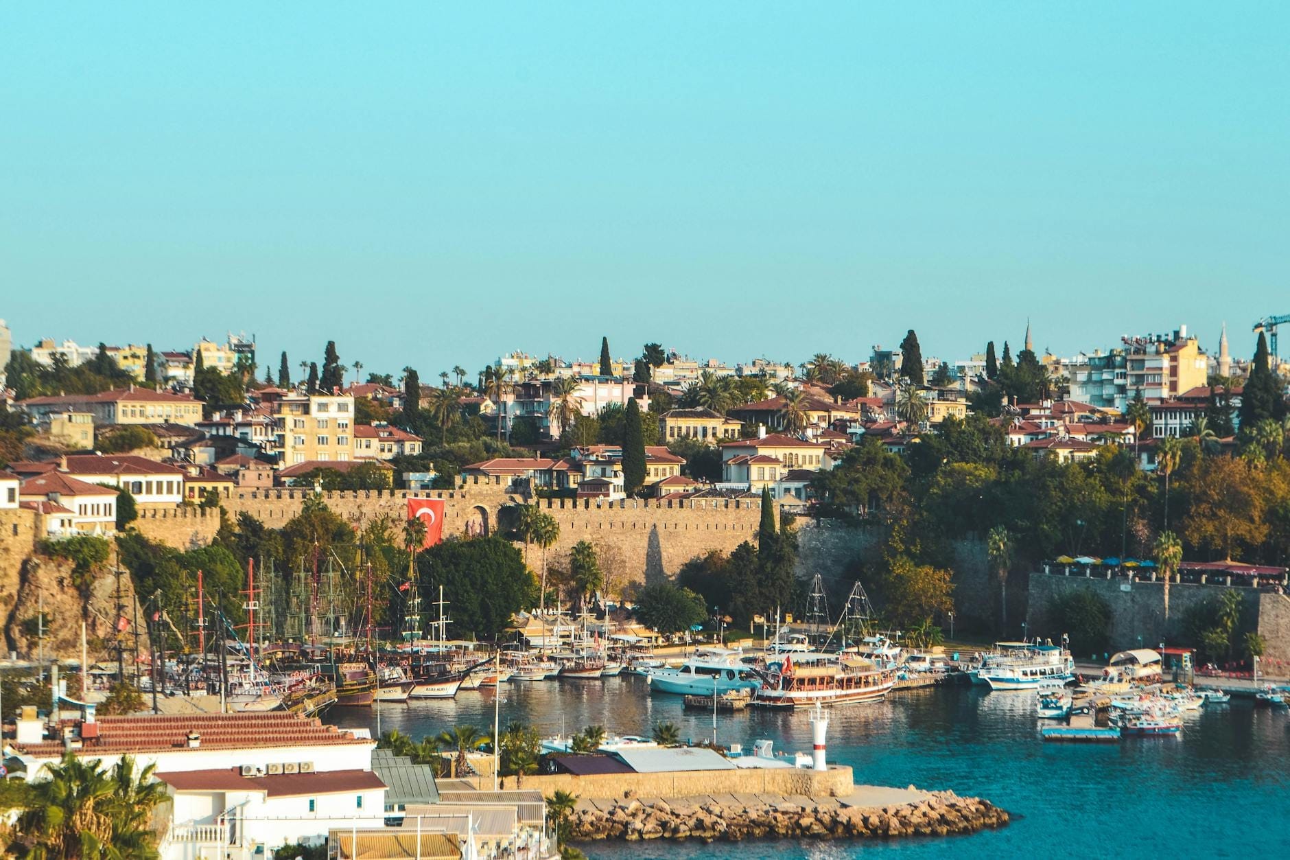 Vista panorâmica do porto da Cidade Velha de Antalya com muralhas históricas, barcos coloridos em águas azuis e arquitetura mediterrânea sob céus limpos.