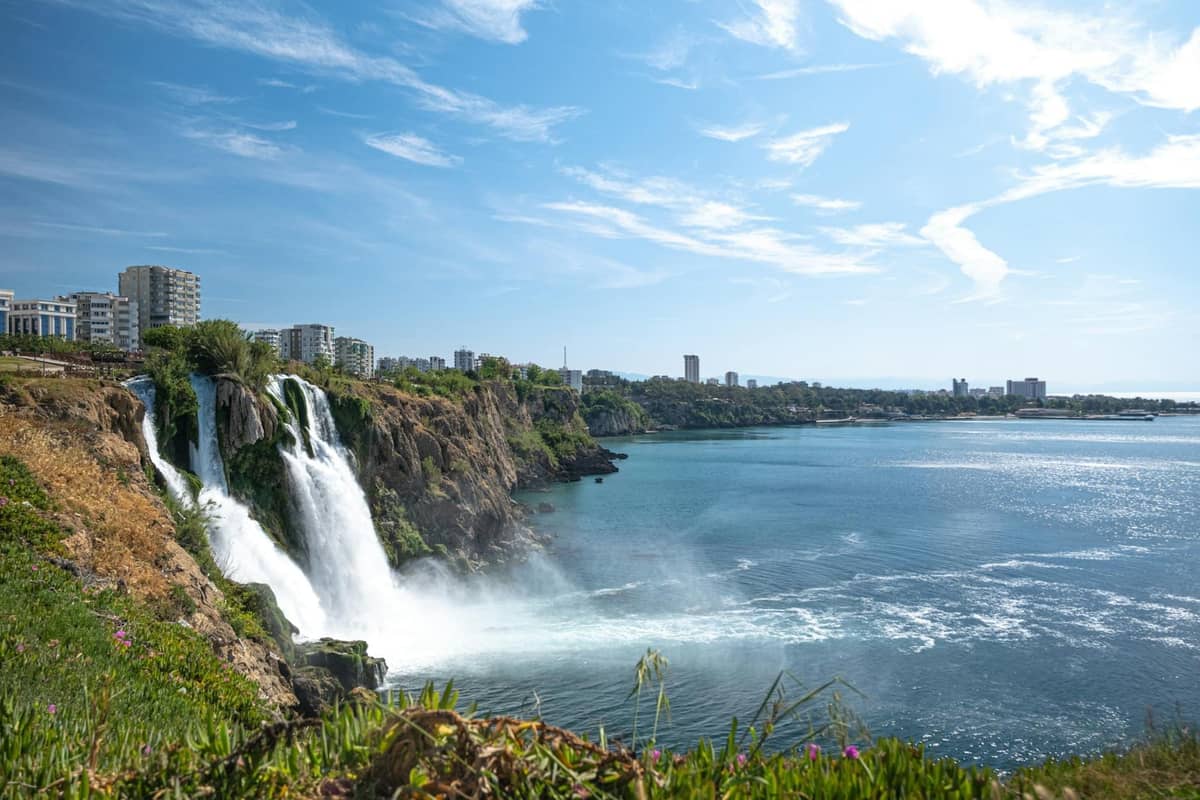 Wide view of Düden Waterfalls cascading into the Mediterranean with city buildings and clear, blue sky in Antalya.