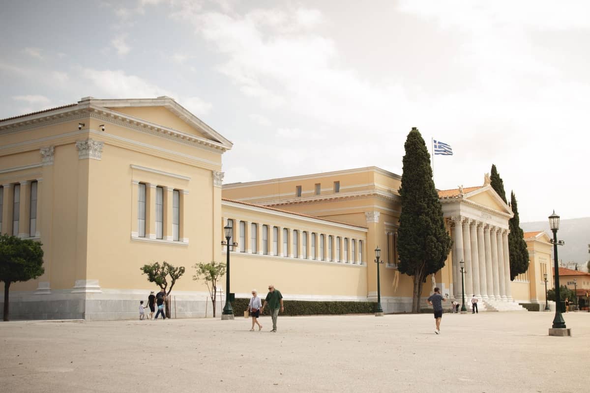 Neoklassizistisches Museumsgebäude mit gelben Wänden, hohen Säulen, Menschen, die draußen spazieren, und einer griechischen Flagge auf dem Dach in Athen.