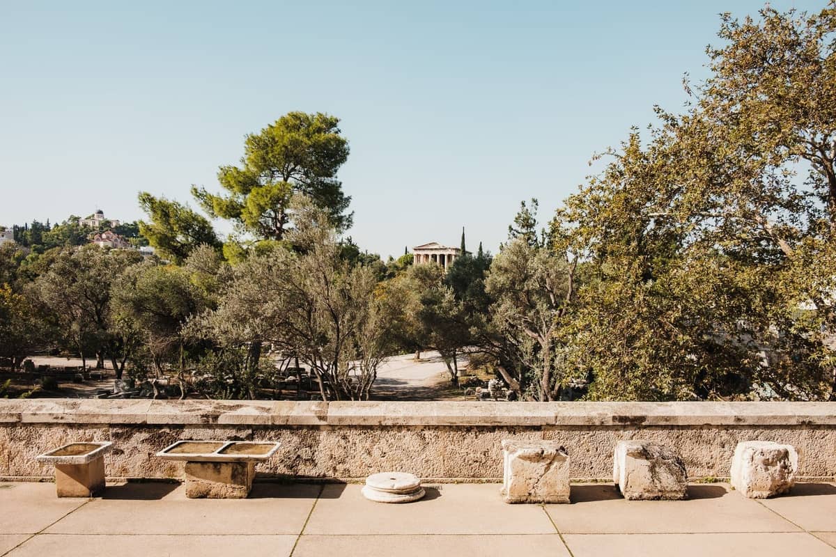 Parkähnliche Anlage in Athen mit antiken Steinsitzen und üppigen grünen Bäumen, mit einem klassischen Tempel in der Ferne unter einem klaren Himmel.