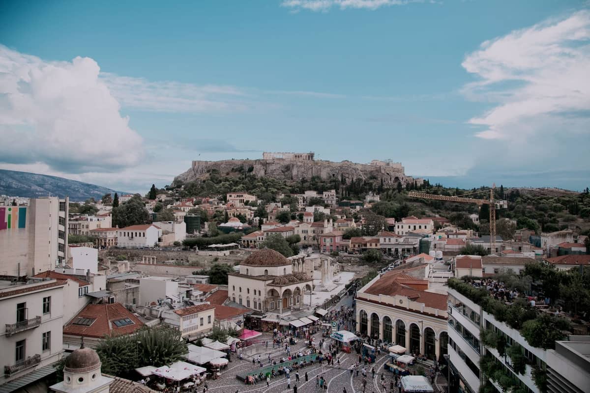 Ein weiter Blick auf Athen mit dem belebten Monastiraki-Platz voller Menschen, Märkten, historischen Gebäuden und der Akropolis im Hintergrund unter einem blauen Himmel.