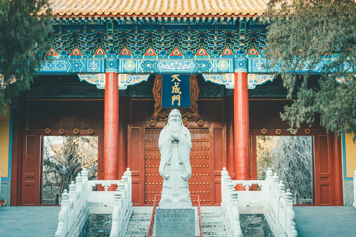 Vorderansicht des Tempeleingangs in Beijing mit roten Säulen, einer großen Konfuzius-Statue, Steinstufen und kunstvollen blau-goldenen Verzierungen.
