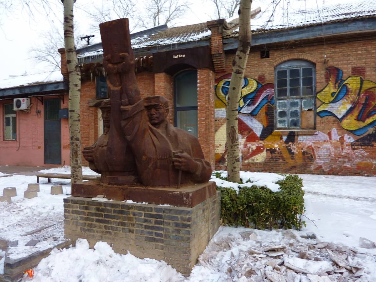Skulptur im Freien mit dem ikonischen Industriegebäude aus roten Ziegeln und bunten Graffiti im 798 Art District in Peking, umgeben von Schnee und kahlen Bäumen.