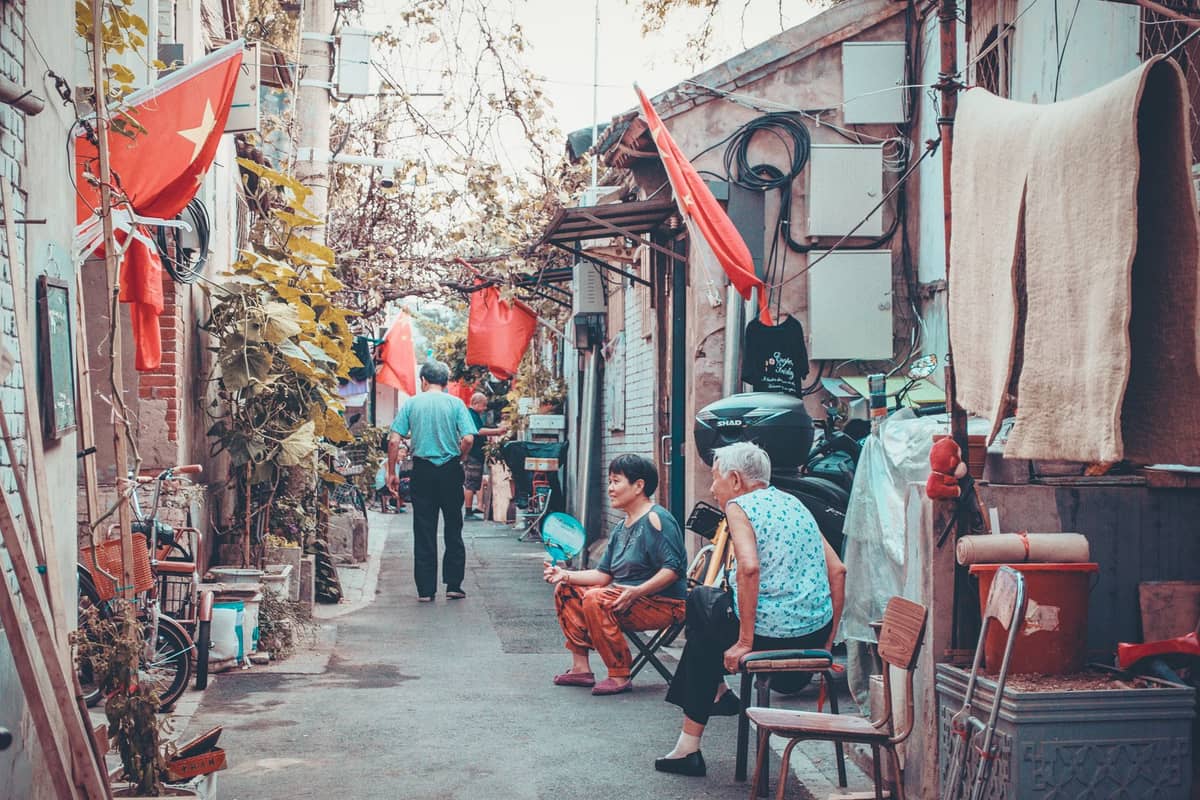 Menschen, die in einer engen Hutong-Gasse in Beijing sitzen und gehen, mit chinesischen Flaggen, Pflanzen, Fahrrädern und alltäglichem Leben.
