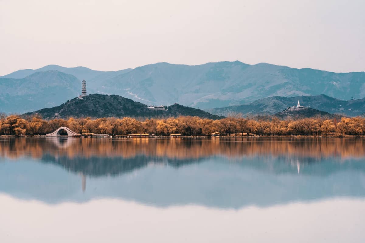 Weiter Blick auf den See des Sommerpalastes in Beijing, mit Bogenbrücke, Pagode, Tempel und herbstlich gefärbten Bäumen, die sich im ruhigen Wasser spiegeln.
