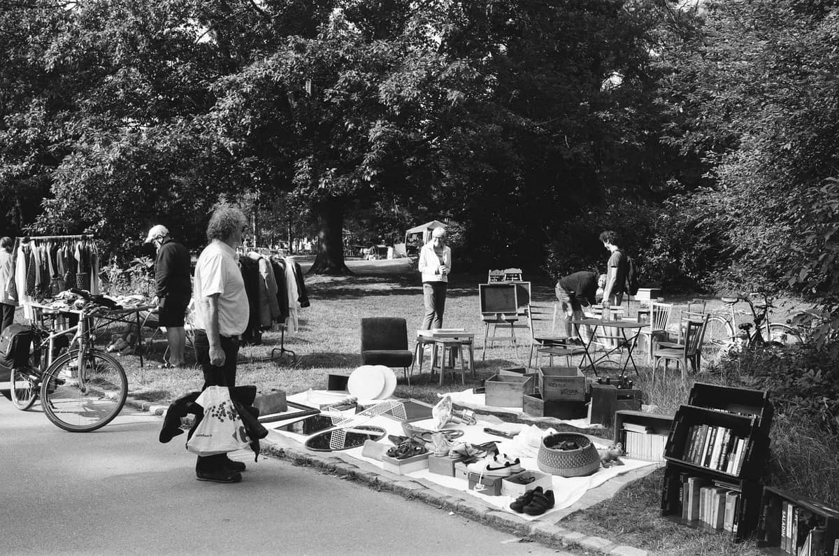 Foto en blanco y negro de personas curioseando artículos en un pequeño mercadillo al aire libre en un parque arbolado, con árboles al fondo.
