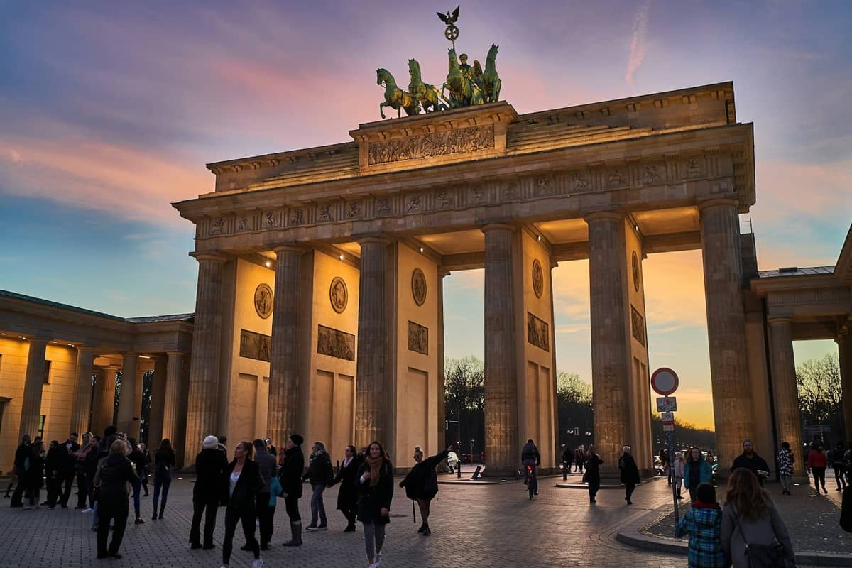 Multitudes de personas caminando frente a la iluminada Puerta de Brandeburgo en Berlín al atardecer, con un colorido cielo sobre ellas.