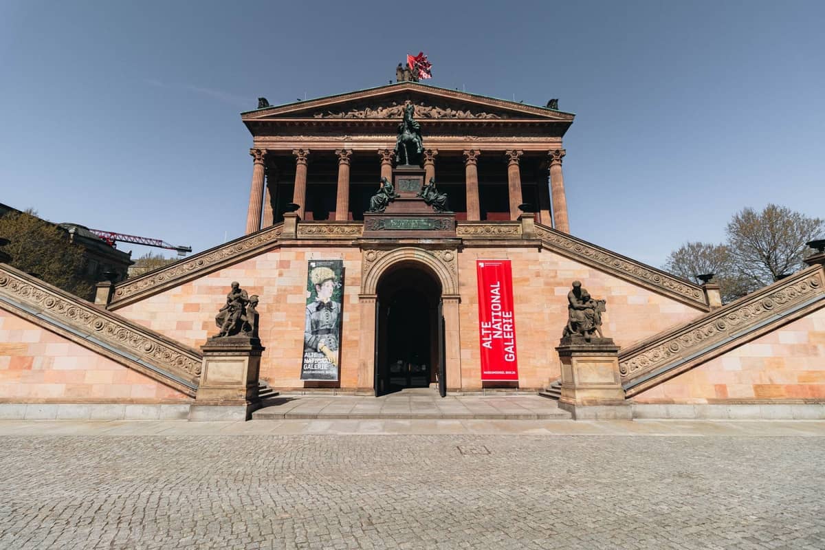 Gran entrada de un museo de Berlín con amplias escaleras de piedra, estatuas, columnas y pancartas con el texto Alte Nationalgalerie. Cielo despejado sobre el edificio histórico.