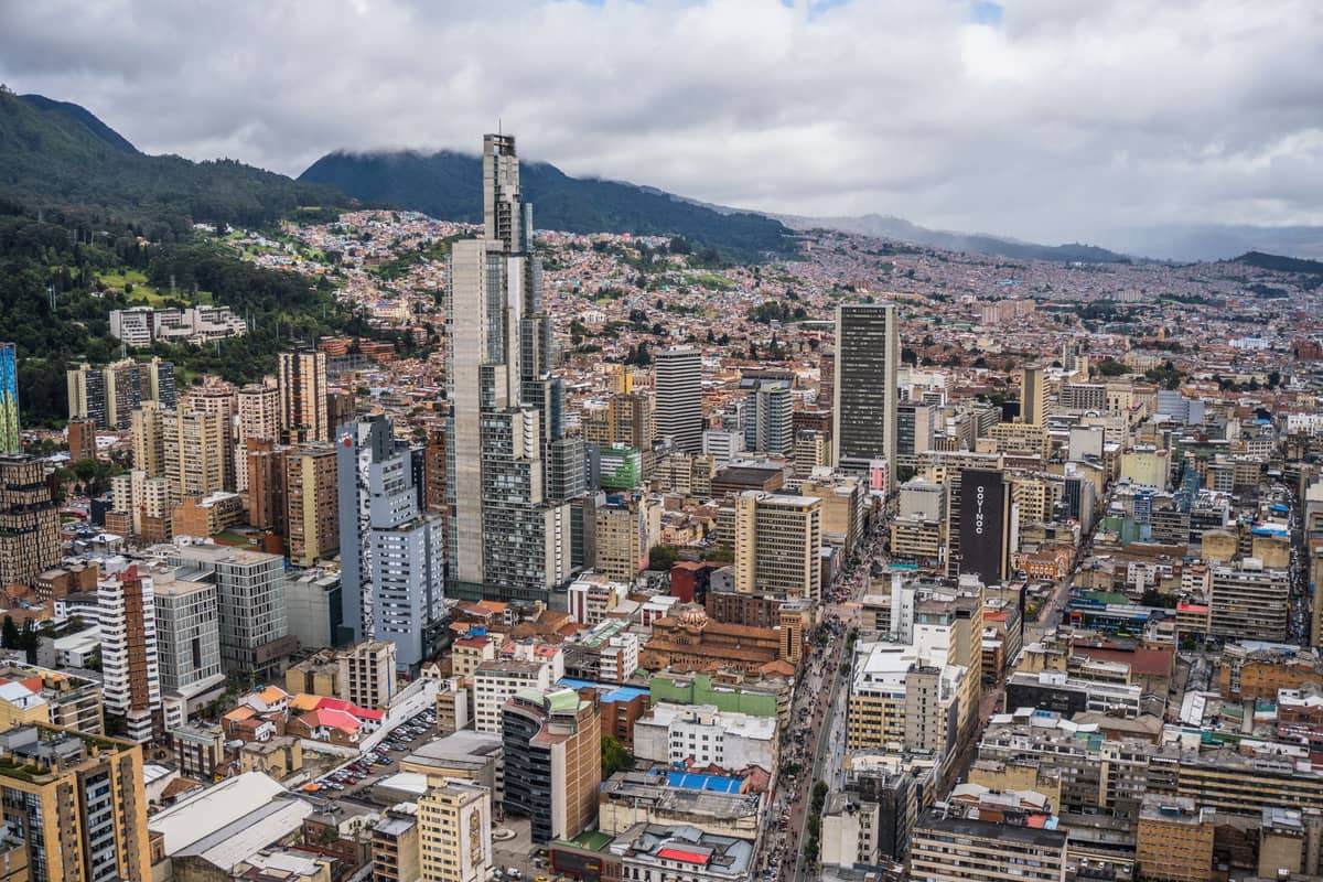 A wide landscape view of downtown Bogotá showing the skyscrapers of the city center with the green Andes mountains in the background.