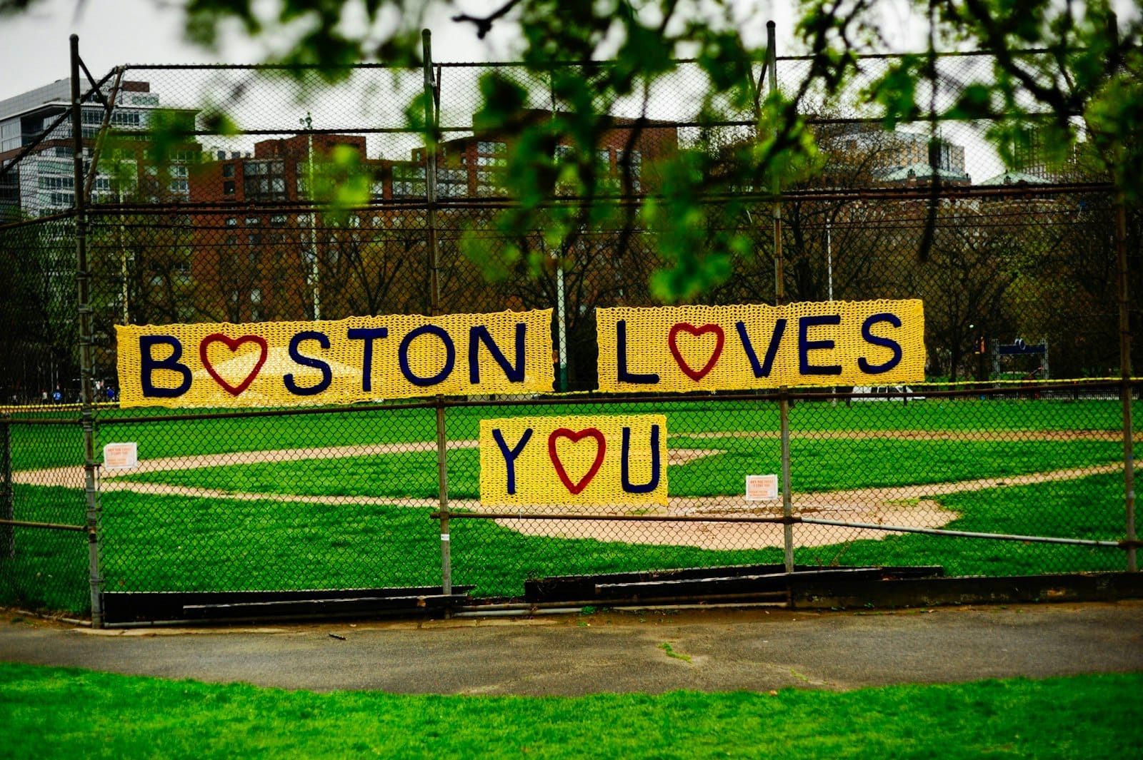 Campo de beisebol do Boston Common com uma cerca de arame decorada com uma placa amarela feita à mão com os dizeres 'Boston Loves You' em um dia nublado.