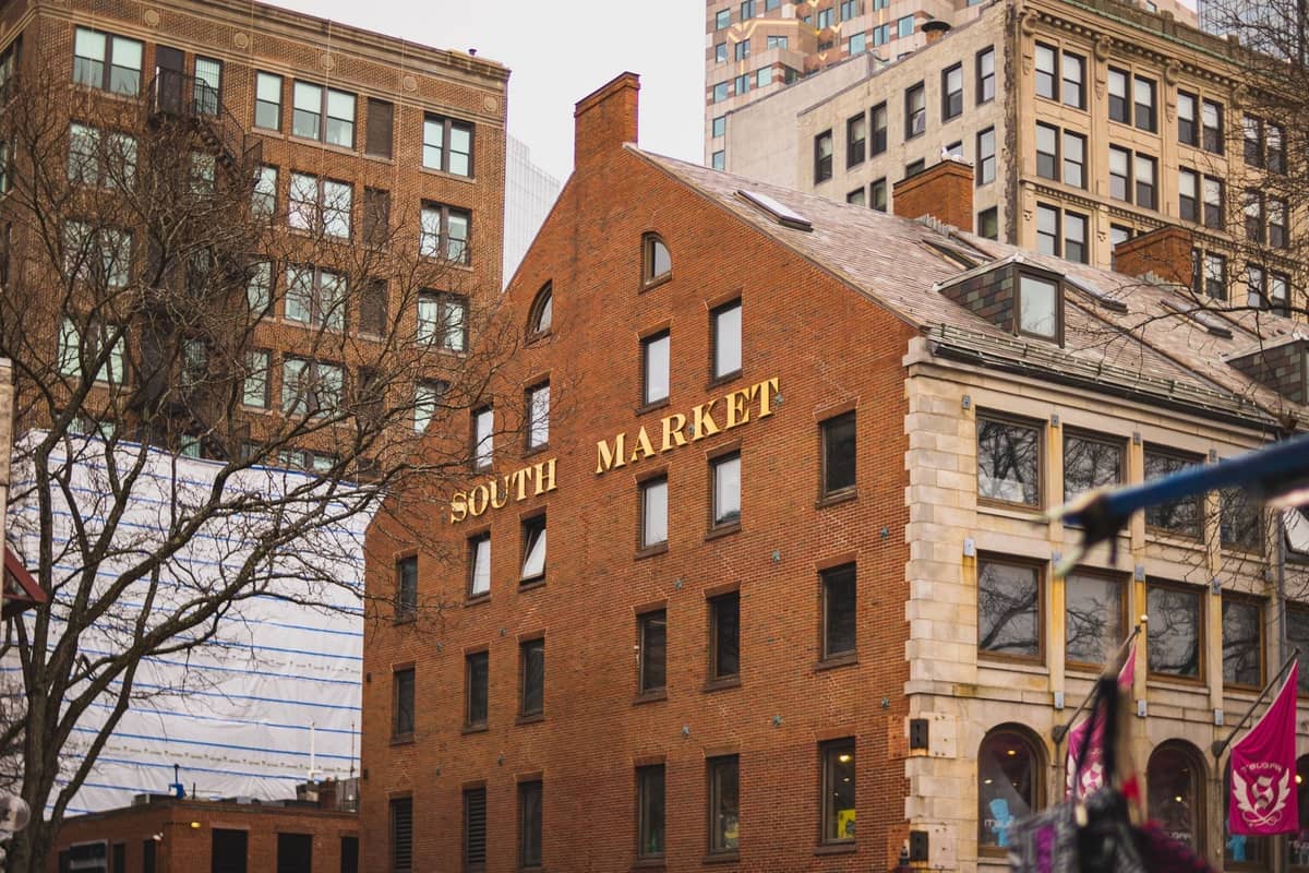 Ceglany budynek z dużymi złotymi literami tworzącymi napis 'South Market' pośród architektury śródmieścia Bostonu w pochmurny dzień.