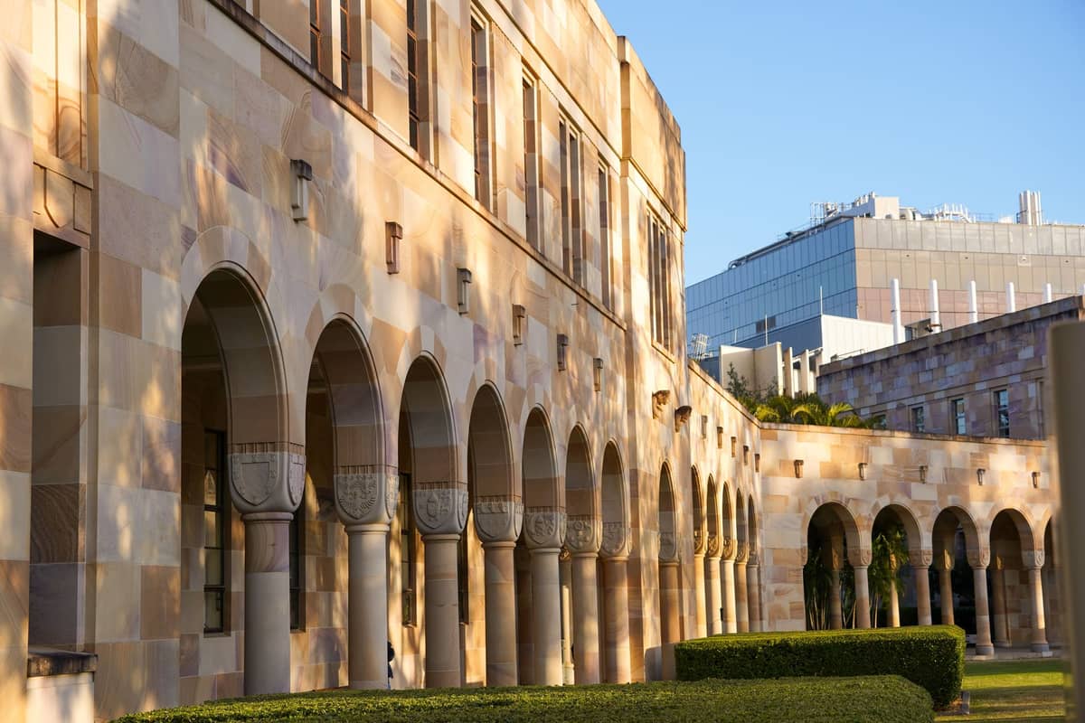 Arcada de arenisca y arquitectura patrimonial de los claustros de la University of Queensland con setos bien cuidados en Brisbane bajo la luz del sol de última hora de la tarde.