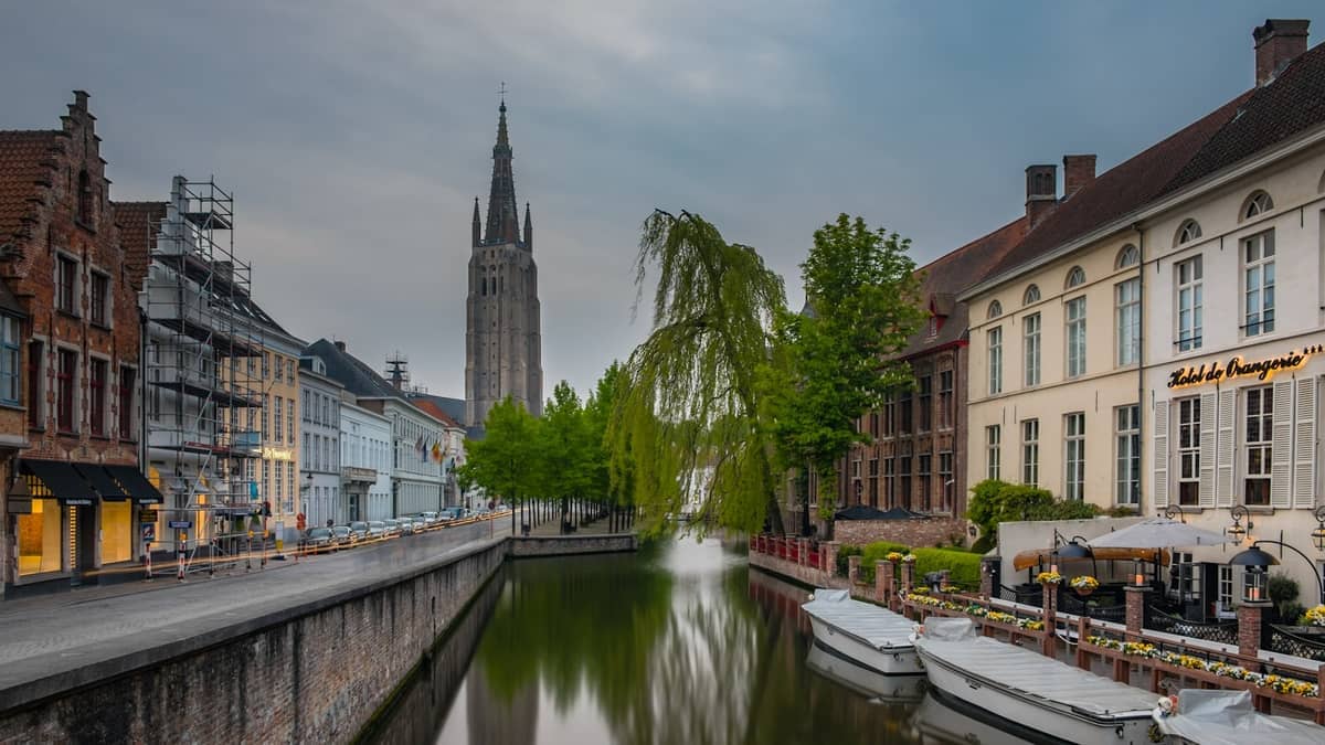 Scenic view of Bruges canal with boats, historic buildings, Hotel de Orangerie, and the Church of Our Lady tower at dusk.