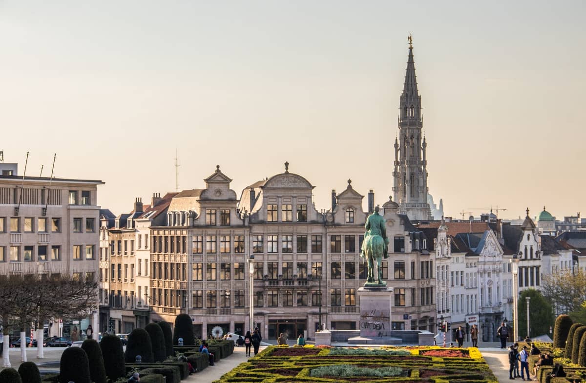 Vista panorâmica do jardim do Mont des Arts com estátua, edifícios históricos e a torre da Câmara Municipal de Bruxelas ao fundo ao pôr do sol.