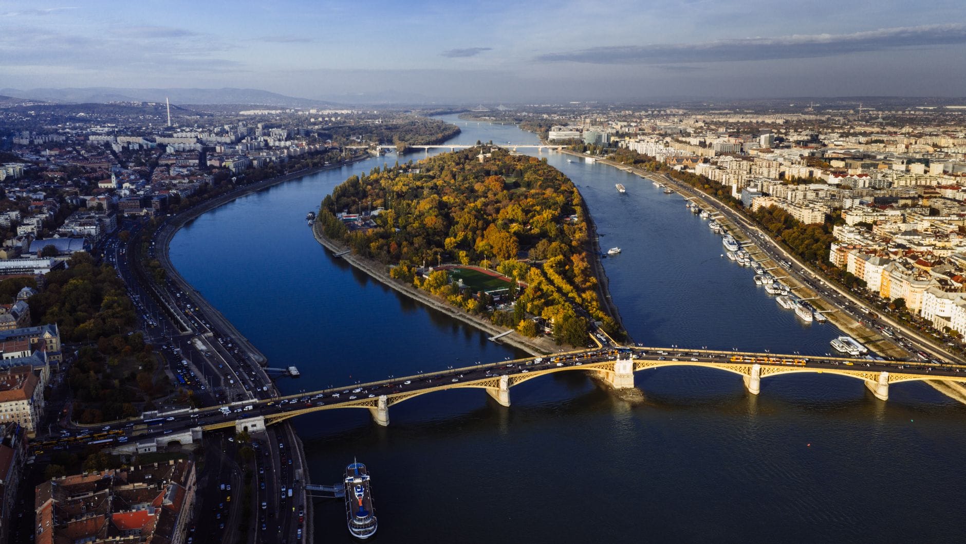 Luftaufnahme der Margareteninsel, einem großen Grünflächenbereich inmitten der Donau im Zentrum von Budapest, mit einer angrenzenden Brücke und Stadtsilhouette.