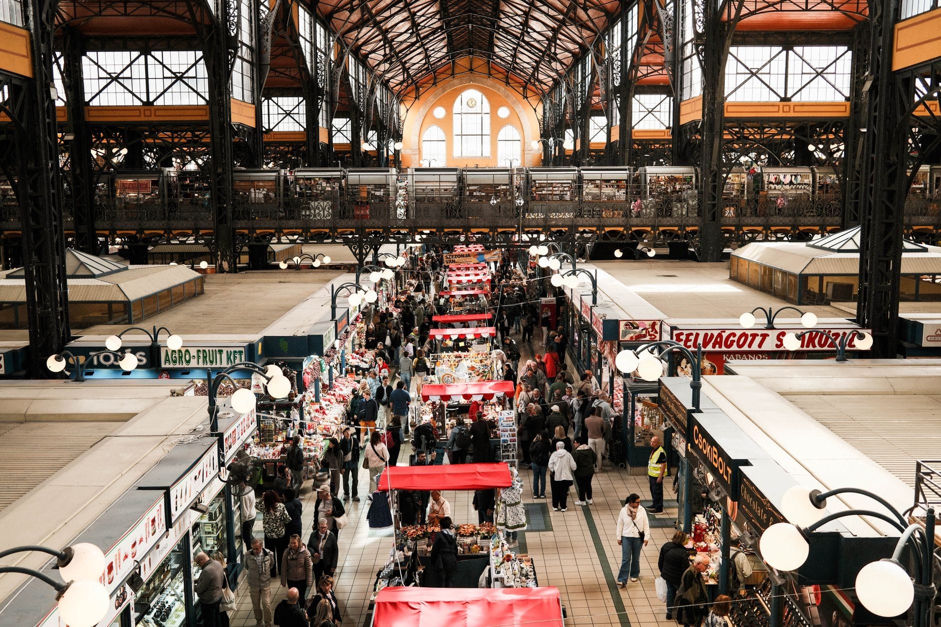 Belebte überdachte Markthalle in Budapest mit Menschenmassen zwischen den Ständen, hohem Gewölbedach und Ständen mit Lebensmitteln und Waren.