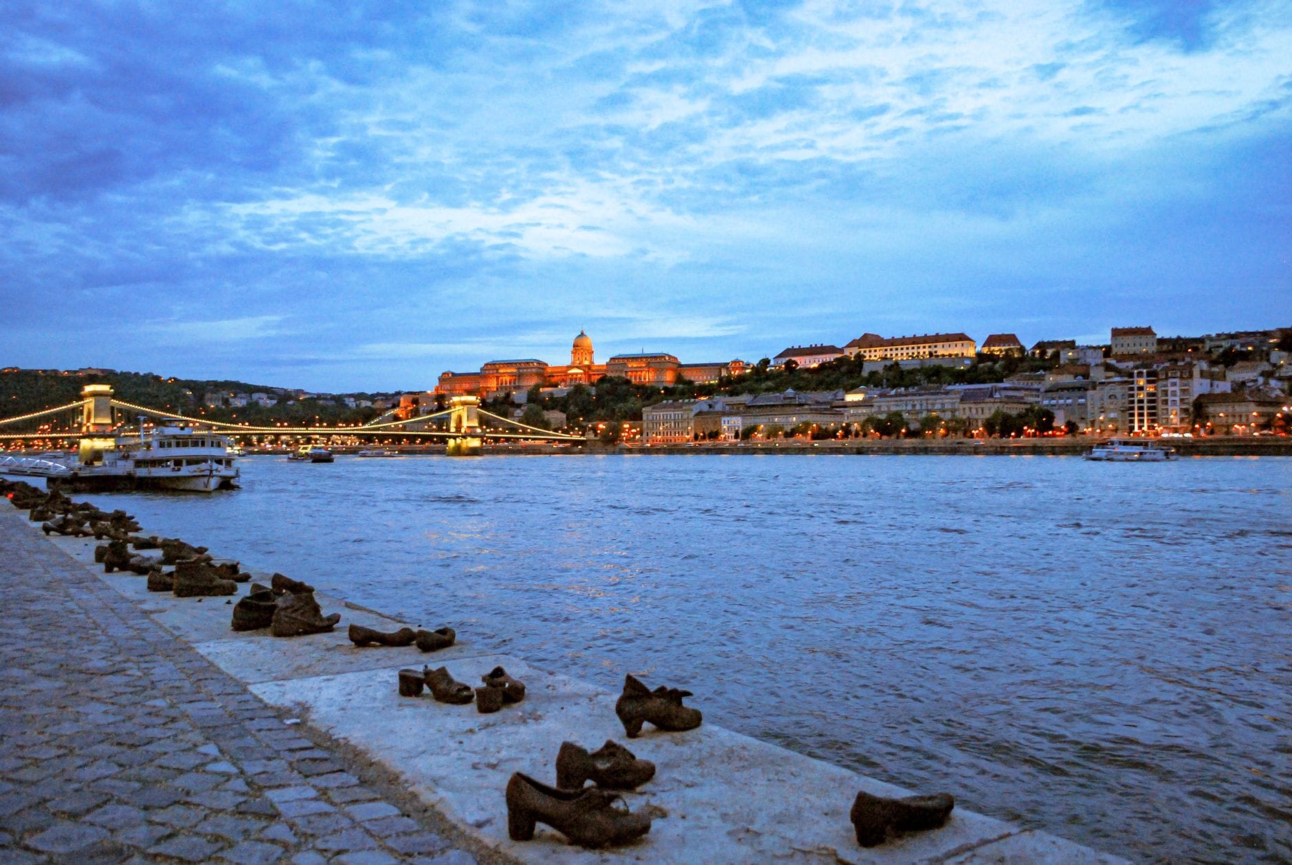 Schuhe-am-Donauufer-Mahnmal mit weitem Blick auf das Flussufer, ikonische Stadtsilhouette von Budapest und die Kettenbrücke in der Abenddämmerung im Hintergrund.