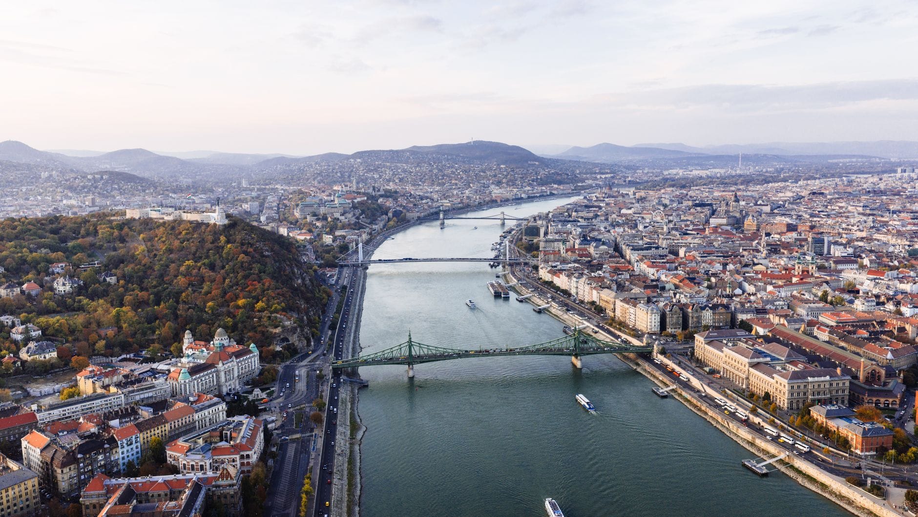 Eine weitläufige Luftaufnahme von Budapest mit der Donau, mehreren Brücken, Gebäuden am Flussufer und den Budaer Hügeln mit ikonischen Stadtpanoramen.