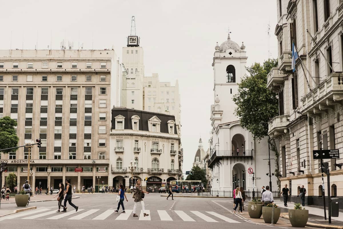 ผู้คนข้ามถนนที่ Plaza de Mayo ในบัวโนสไอเรส ล้อมรอบด้วยอาคารอาณานิคมสีขาวที่มีประวัติศาสตร์และสถาปัตยกรรมสมัยใหม่ใต้ท้องฟ้าที่มีเมฆครึ้ม