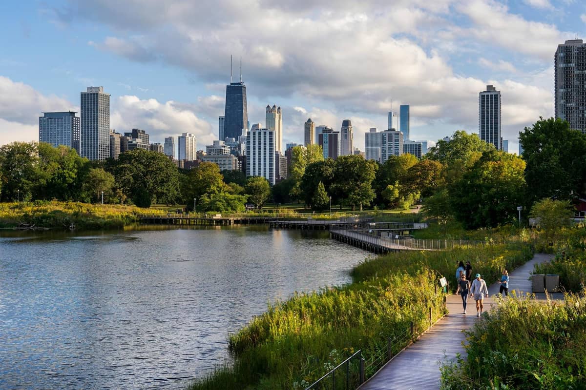 Skyline von Chicago, von einem Parkgelände am Seeufer aus gesehen, mit Menschen, die auf einem von Grün und Wasser umgebenen Steg spazieren.