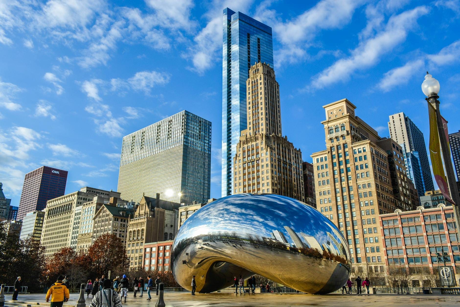 A escultura Cloud Gate, conhecida como The Bean, cercada de pessoas com o skyline de Chicago ao fundo em um dia ensolarado.