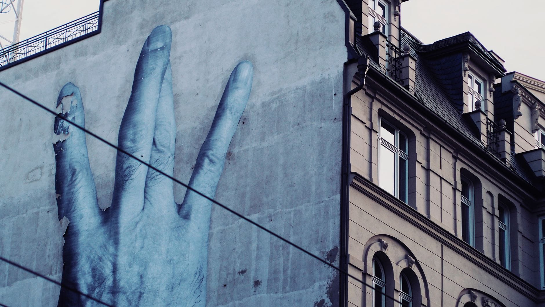 จิตรกรรมฝาผนังขนาดใหญ่รูปมือสีน้ำเงินบนผนังอาคารในย่านเมือง พร้อมสายไฟรถรางที่มองเห็นได้และสถาปัตยกรรมดั้งเดิมของ Köln