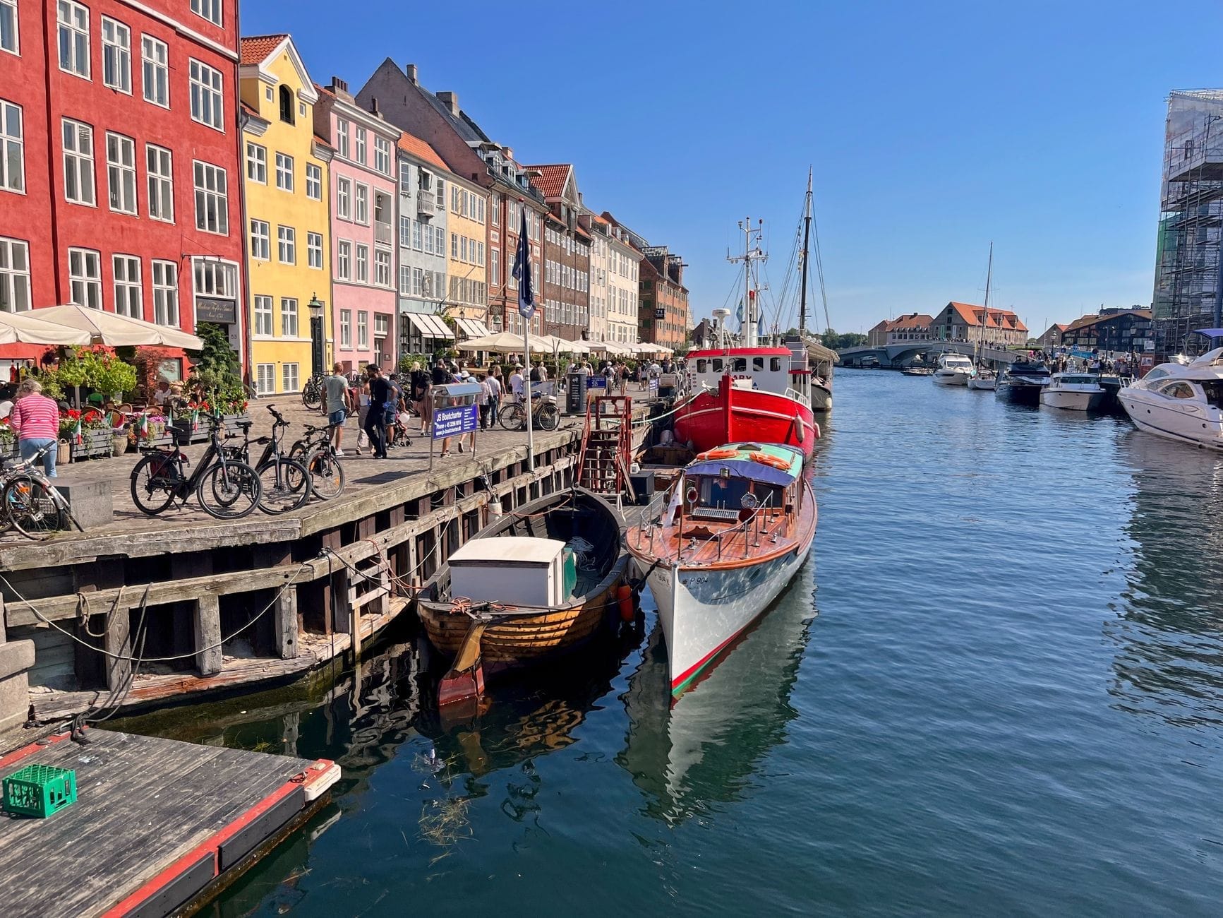 Casas históricas coloridas e cafés movimentados ao longo do canal Nyhavn, em Copenhague, com barcos atracados e pessoas aproveitando um dia de sol.