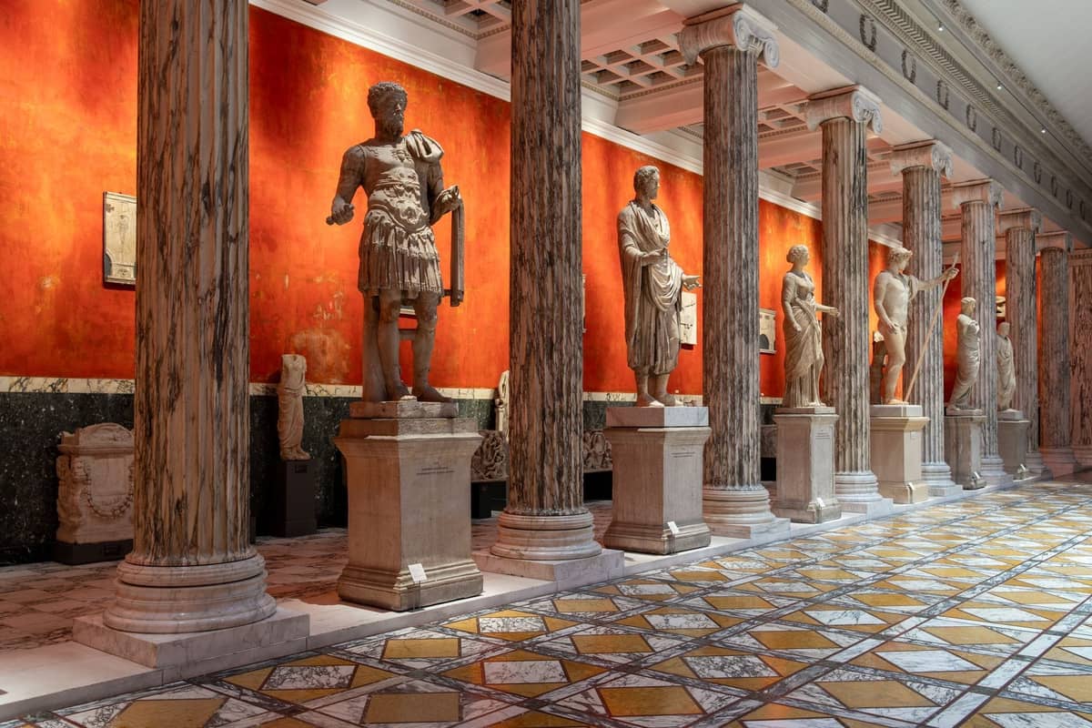 Sala de un museo de arte con altas columnas de mármol y estatuas de estilo romano frente a una llamativa pared roja, probablemente en Copenhagen.