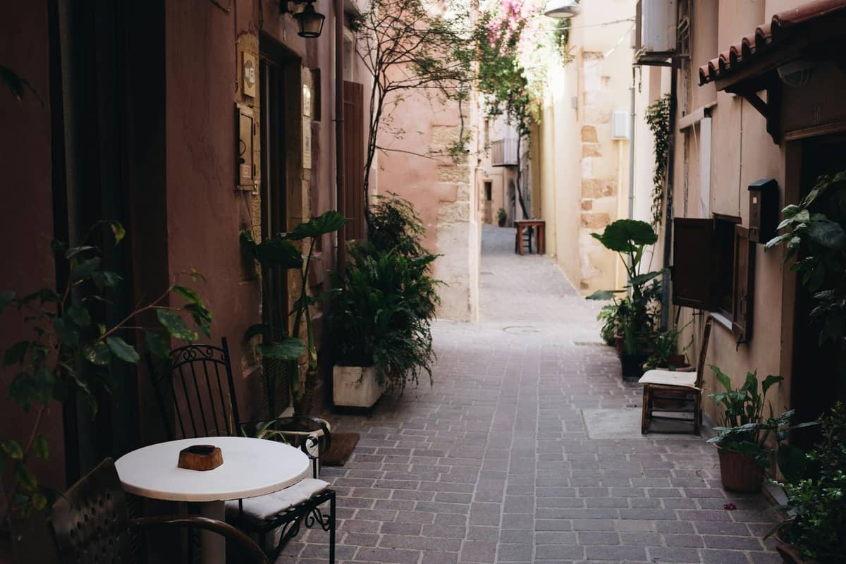 Enge Fußgängergasse in Kretas historischer Altstadt mit Topfpflanzen, einem Cafétisch und sonnenbeschienenen Steingebäuden.