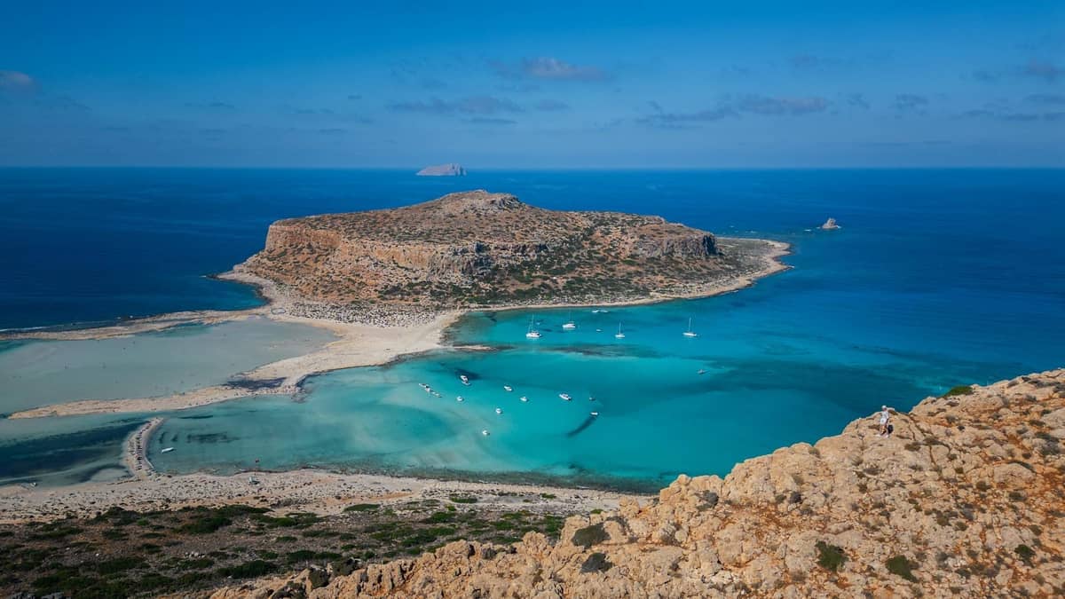 Panoramablick auf den Balos Beach auf Kreta mit türkisfarbenem Wasser, sandigen Lagunen, felsiger Küste und in der Bucht ankernden Booten unter klarem Himmel.