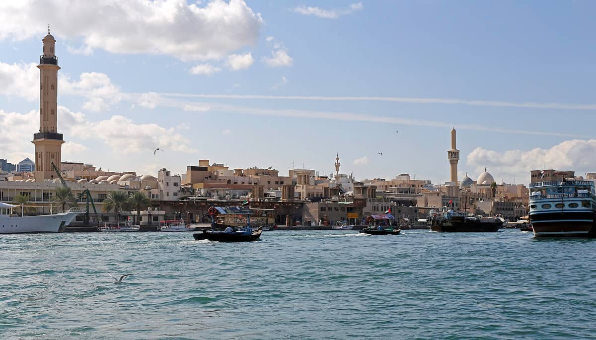 Blick auf den Dubai Creek mit traditionellen Abras und historischen Gebäuden, darunter Windtürme und Minarette, unter einem teils bewölkten blauen Himmel.