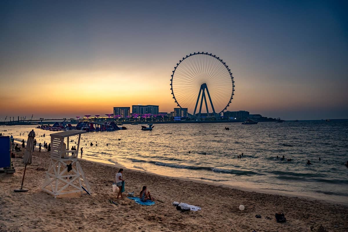 JBR Beach bei Sonnenuntergang mit Menschen im Sand, einem Rettungsschwimmerstuhl und dem Riesenrad Ain Dubai, das über das Wasser zu sehen ist.