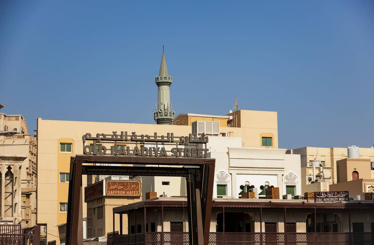 Straßenszene in der Old Baladia Street in Dubai, mit Moschee-Minarett, traditionellen Gebäuden und klarem blauen Himmel.