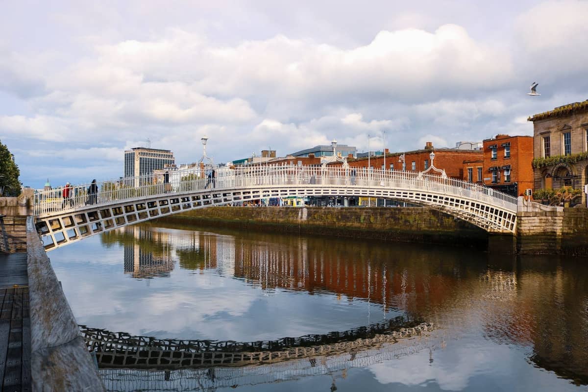 Tagesansicht der Ha'penny Bridge über den River Liffey in Dublin, mit Fußgängern, roten Backsteingebäuden und Spiegelungen im Wasser.