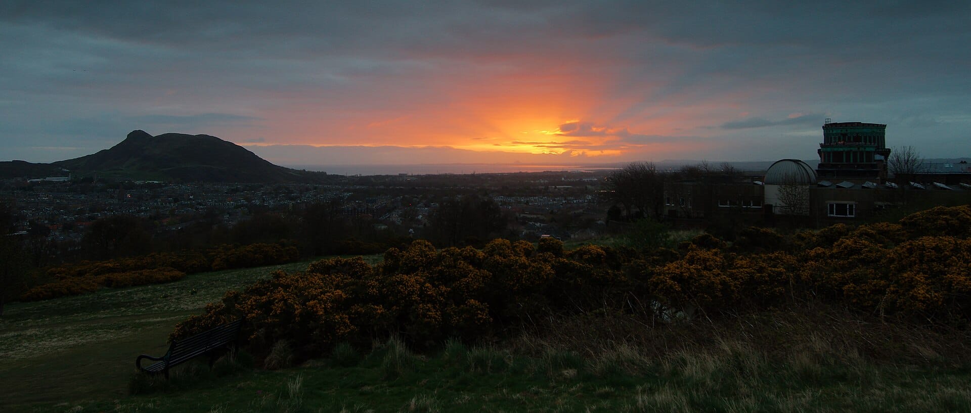 Weite Sonnenuntergangsansicht von Arthur's Seat und den Salisbury Crags über Edinburgh, mit Parkwiesen und einem Observatorium im Vordergrund.