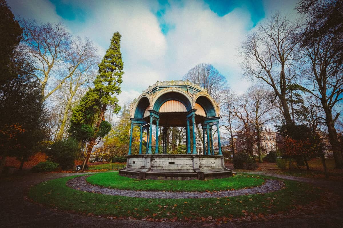 Decorative gazebo surrounded by trees and greenery in a landscaped park in Ghent, on a partly cloudy day.