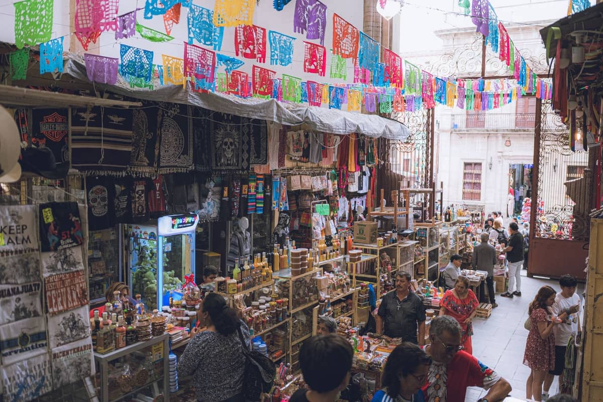 ฉากตลาดในร่มที่มีธง papel picado หลากสีสัน แผงขายงานหัตถกรรม และผู้คนที่กำลังเลือกชมสินค้าในตลาดที่คึกคักของ Guadalajara