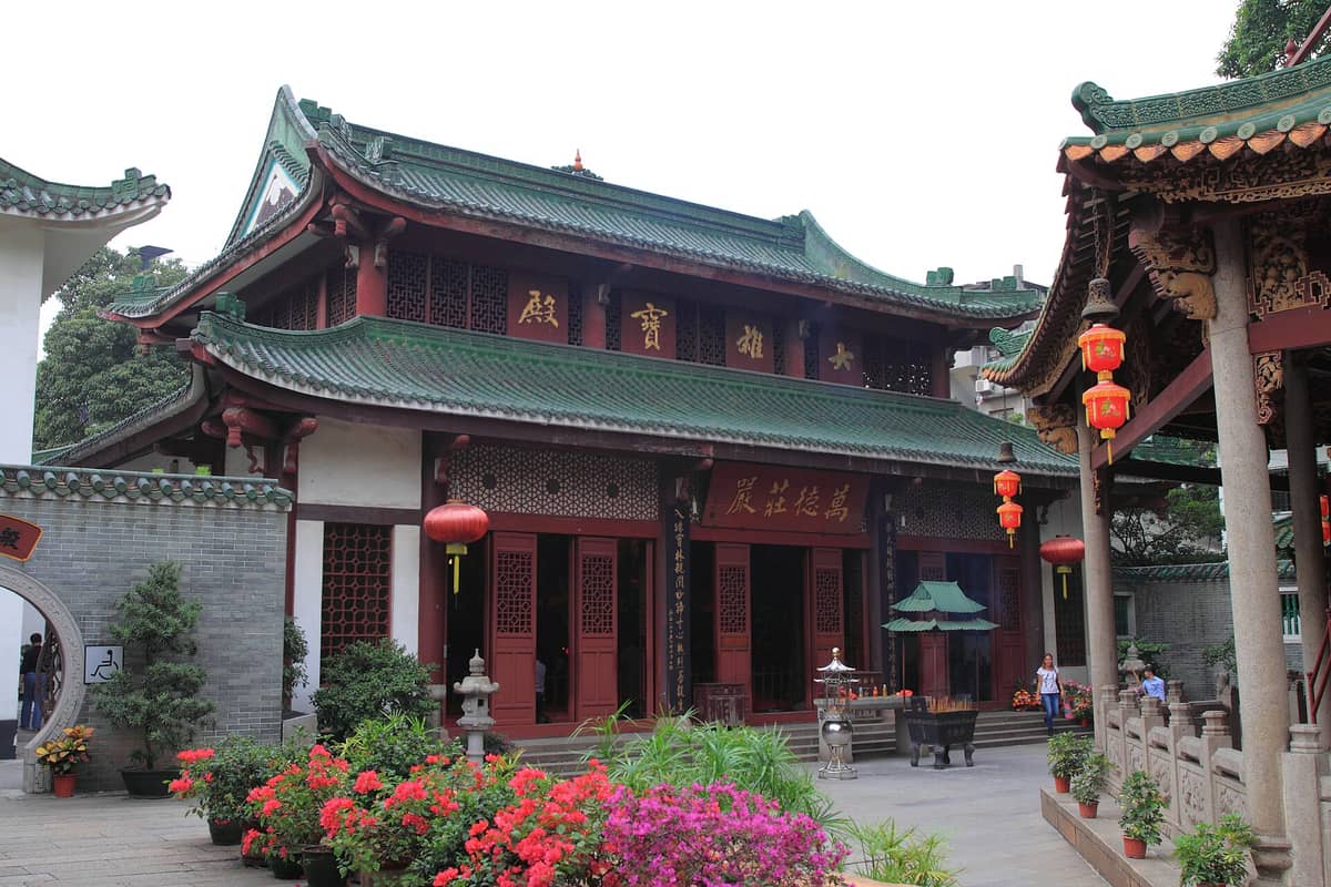 Courtyard of a historic Chinese temple in Guangzhou with green-tiled roofs, red pillars, potted plants, incense burner, and hanging red lanterns.
