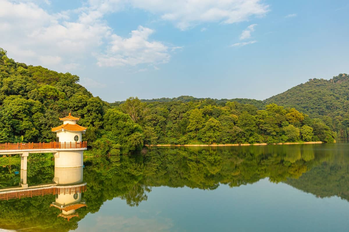Scenic view of a lush green lakeside park with forested hills and a small traditional pavilion on the water in Guangzhou.