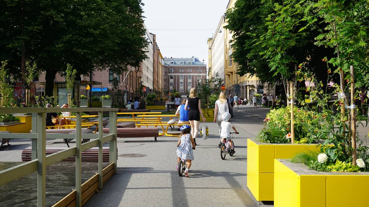 Rua amigável para pedestres no bairro Punavuori de Helsinque com famílias, ciclistas, bancos e vibrantes jardineiras amarelas, destacando um espaço urbano focado no design e atmosfera criativa.
