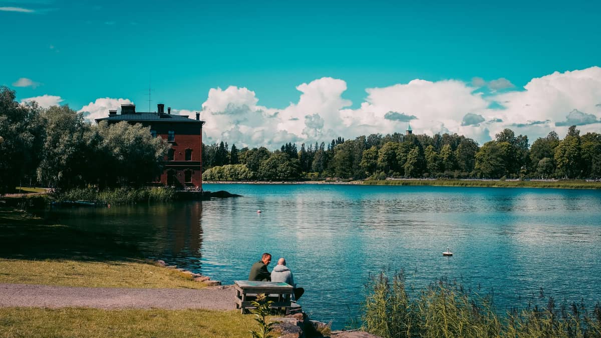 Duas pessoas sentadas num banco junto a uma orla tranquila em Helsinki, com árvores, relva e um edifício de tijolos vermelhos ao fundo.