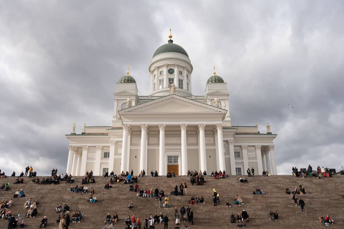 Vista ampla da Catedral de Helsinque com multidões sentadas nos grandes degraus sob um céu nublado dramático.