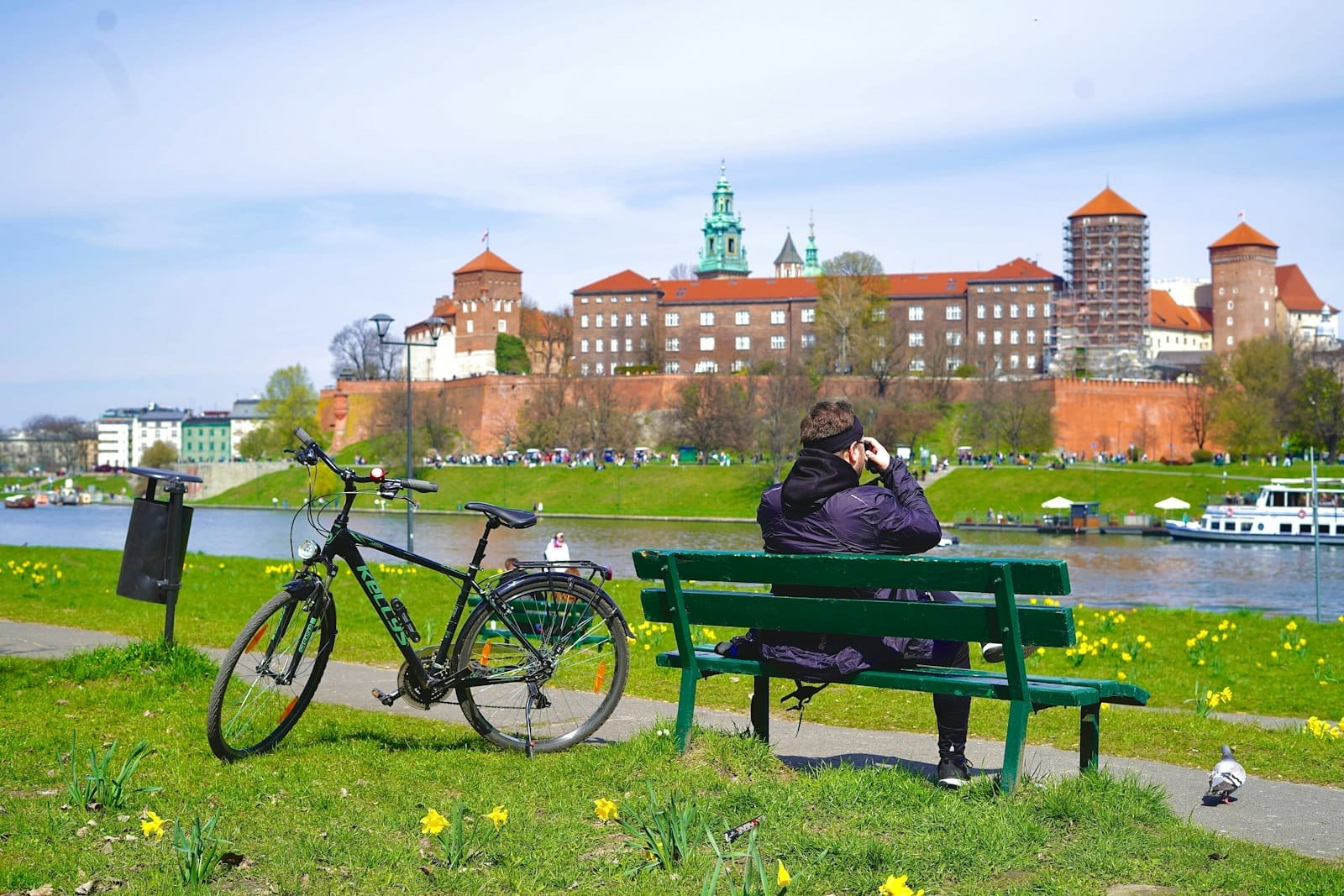 บุคคลนั่งบนม้านั่งสีเขียวพร้อมจักรยานอยู่ใกล้ๆ มองเห็นปราสาท Wawel ริมแม่น้ำ Vistula ในวันที่แดดจ้าที่เมือง Kraków
