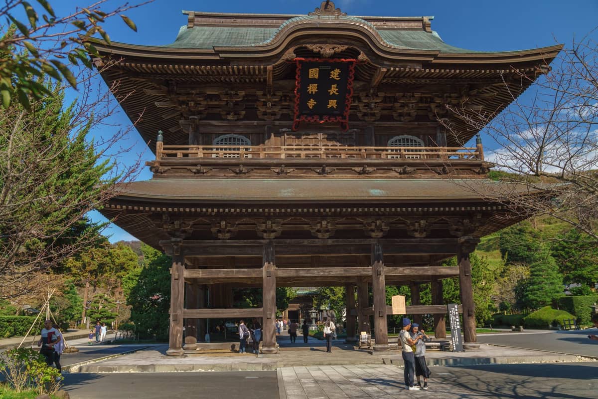 Large wooden temple gate in Kyoto with people walking beneath it, trees and temple grounds in background, sunny day.
