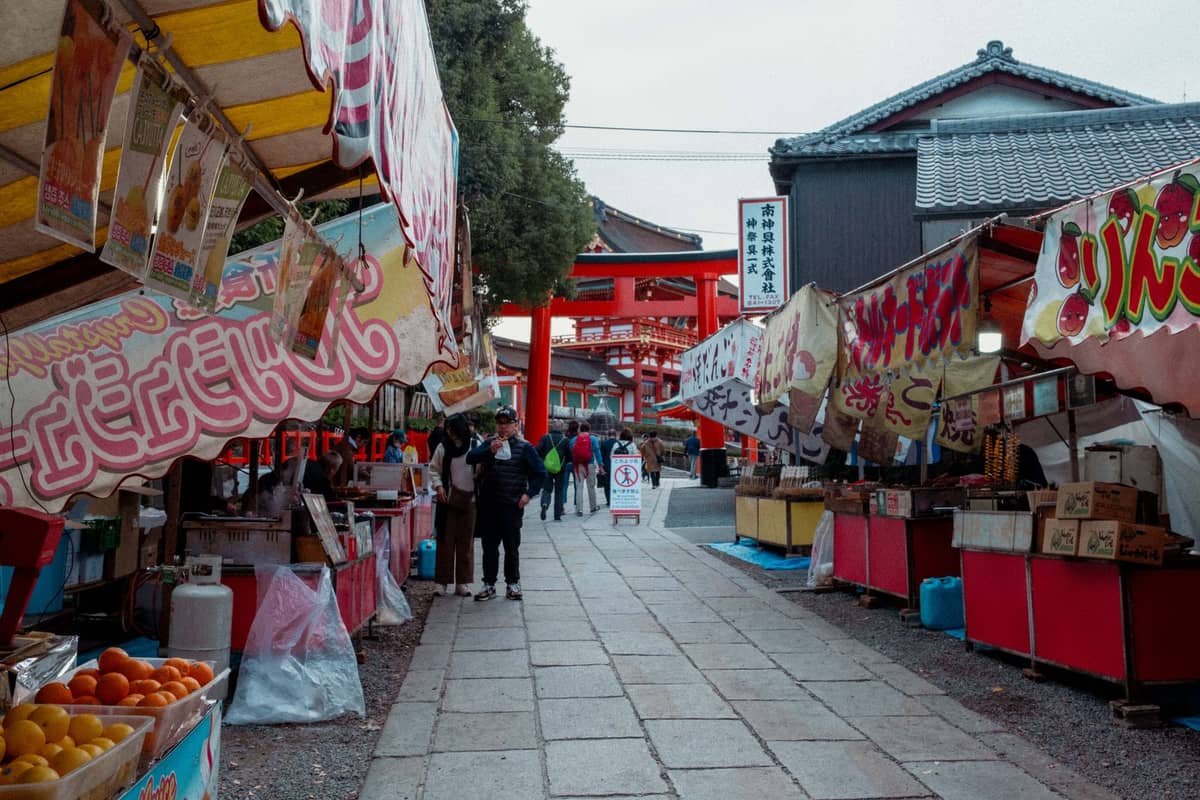 Street scene in Kyoto with food stalls on both sides, people walking, and a red torii gate in the background.