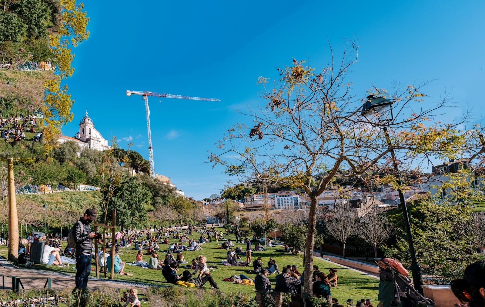 Pessoas relaxando e socializando em uma encosta gramada de parque em Lisboa sob um céu azul claro com edifícios da cidade ao fundo.