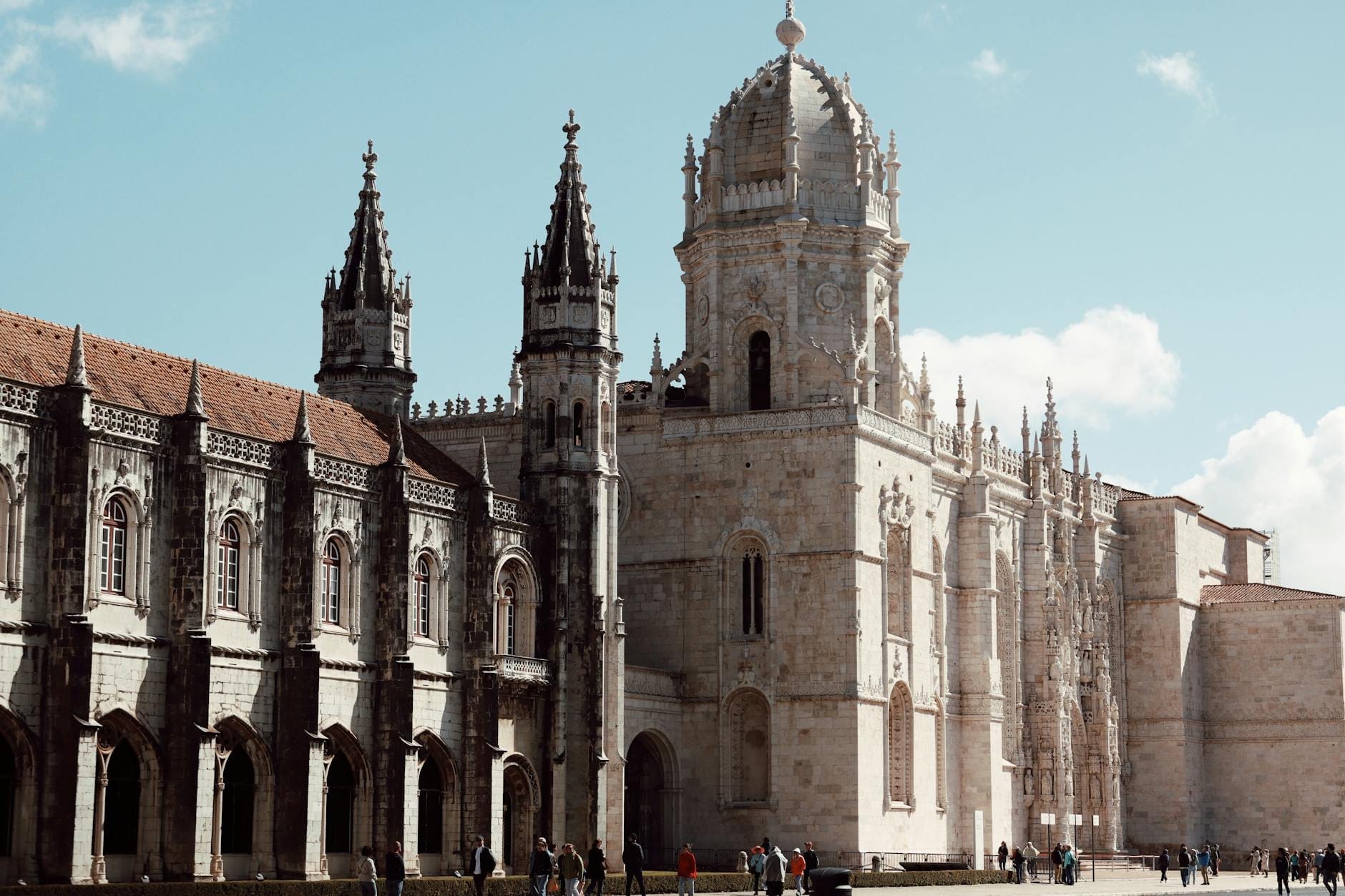 Vista ampla da fachada ornamentada e torres do Mosteiro dos Jerónimos, com pessoas caminhando em primeiro plano sob um céu azul em Lisboa.