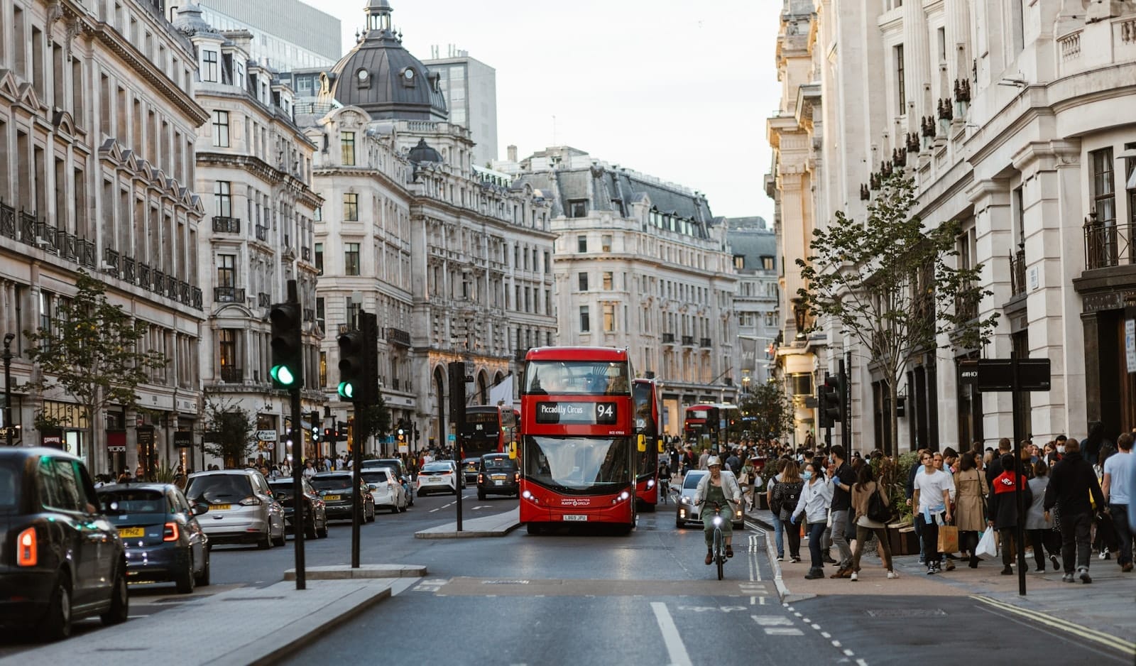 Movimentada cena de rua em Londres perto de Piccadilly Circus com clássicos ônibus vermelhos de dois andares, edifícios históricos, carros e pedestres sob uma suave luz de tarde.